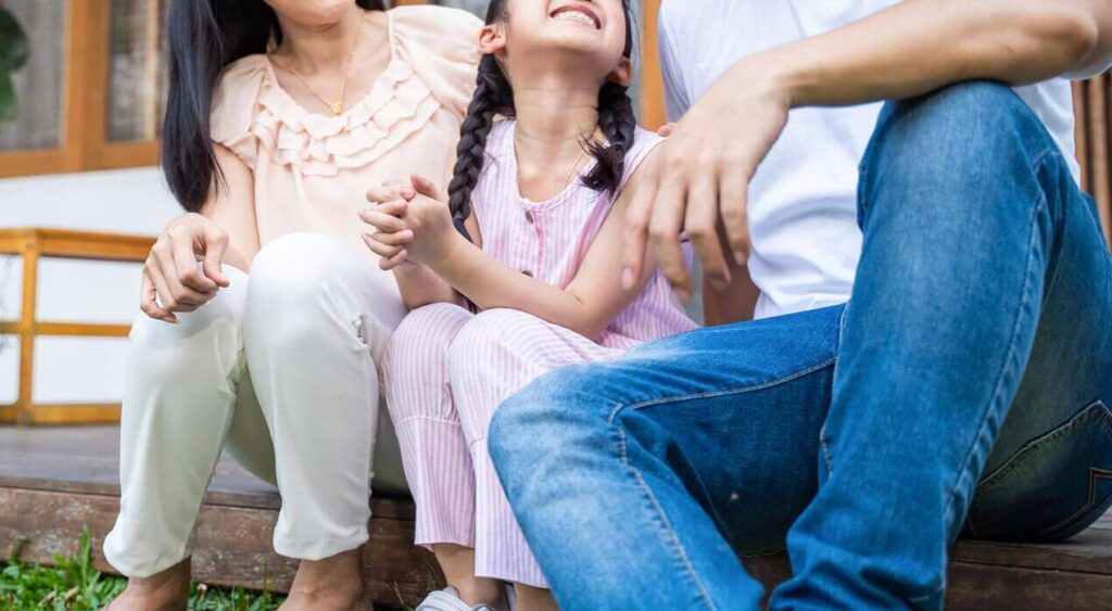 Image of a smiling Asian family siting outside on a step. If your child is struggling to cope with their emotions and behaviors, find support by working with a skilled child therapist in South Jersey to learn mindfulness.