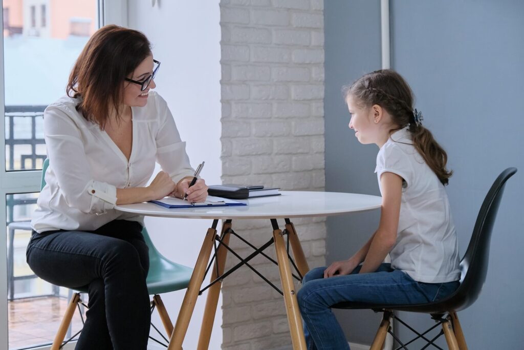 Image of a therapist sitting at a table with a little girl. With therapy for children in South Jersey, you can start seeing positive behaviors in your child as they manage their emotions.