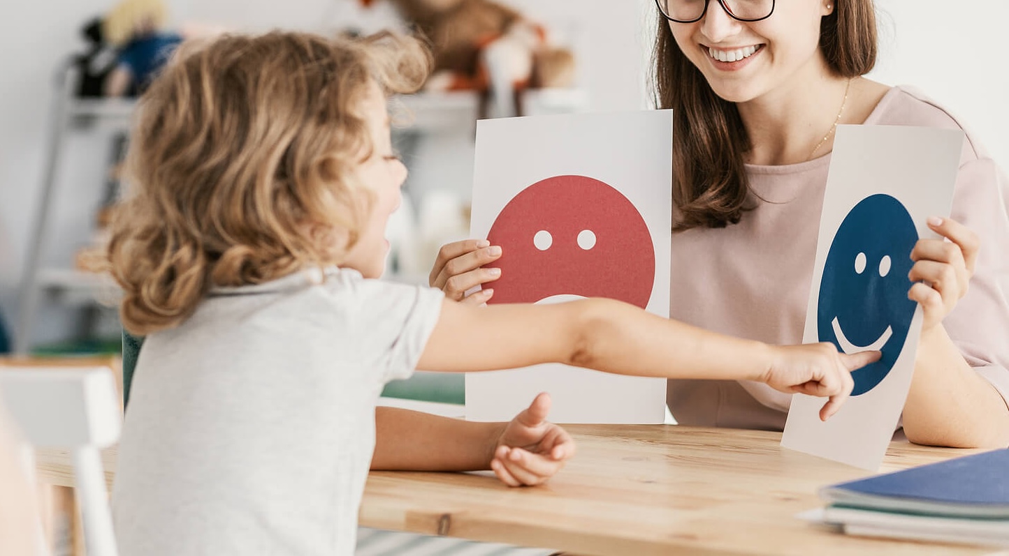 Image of a young boy pointing at face emotions on a paper with a smiling therapist. If your child struggles to manage their unwanted emotions and behaviors, find support with a skilled child therapist in South Jersey to help them better understand themselves.