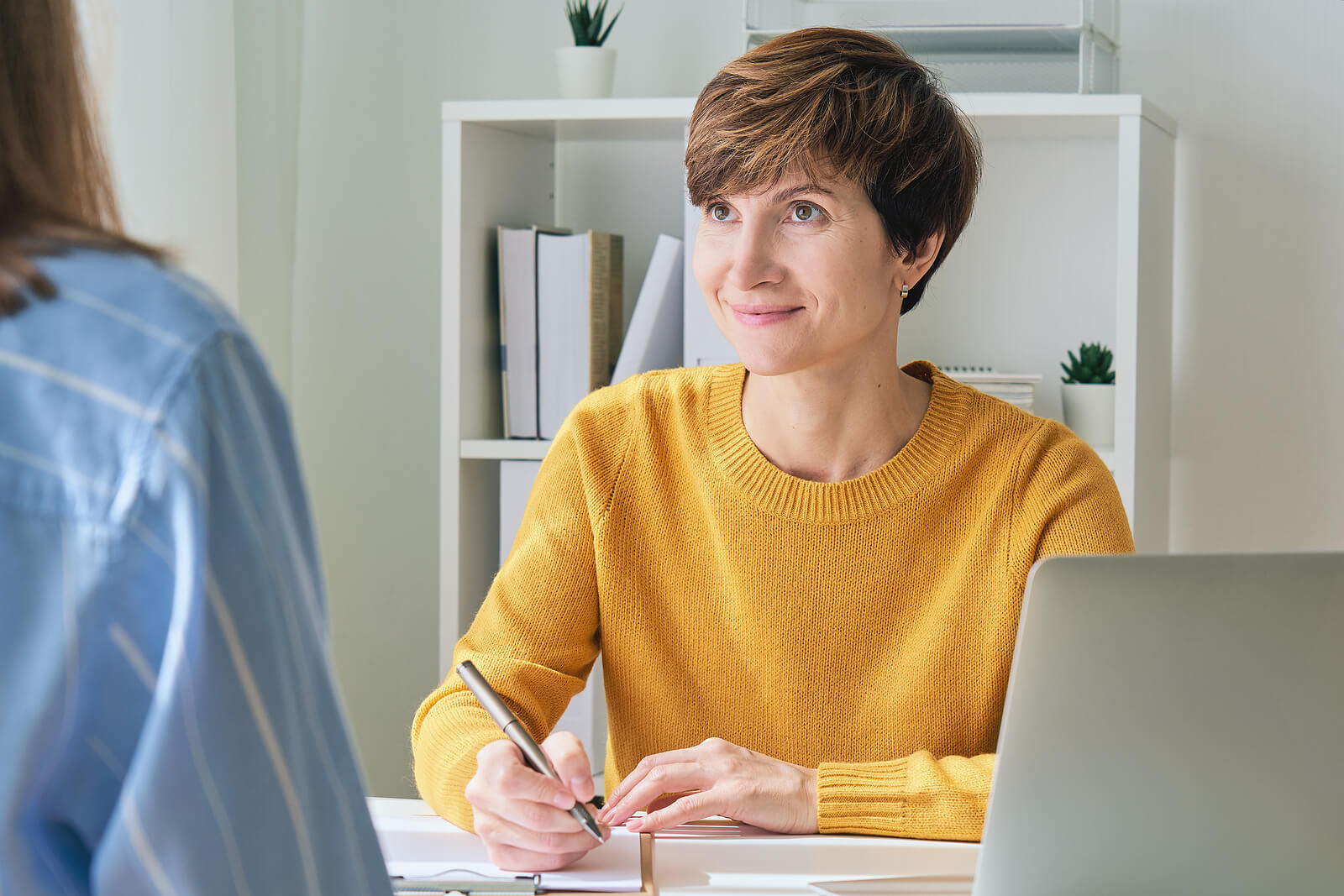 Smiling woman in a yellow sweater holding a pen while listening to another woman talk. Find self-acceptance and learn to cope with your challenges in positive ways with ADHD testing in South Jersey.