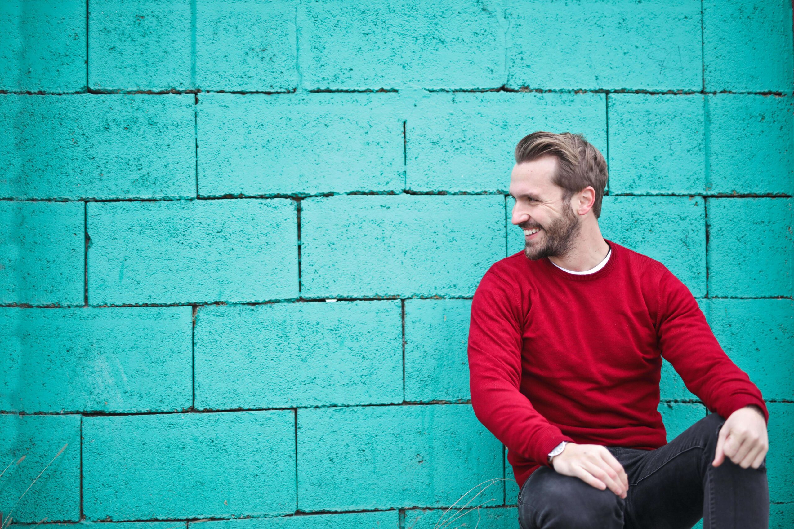 Smiling man wearing a red sweater while sitting in front of a teal brick wall. Celebrate your true self and cope with your challenges in positive ways with the support of a neurodivergent affirming therapist in South Jersey.