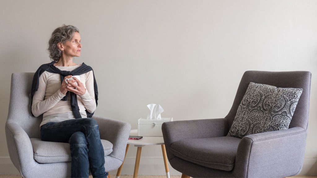 A woman sits alone in an armchair holding a mug of tea, looking thoughtfully toward the empty chair beside her, capturing the quiet adjustment parents face when their child leaves for college in New Jersey