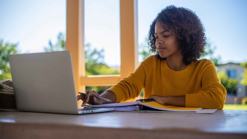 A young woman with curly hair sits at a desk near a sunlit window, writing in a notebook beside her laptop, reflecting the balance of coursework and support that online therapy offers New Jersey college students