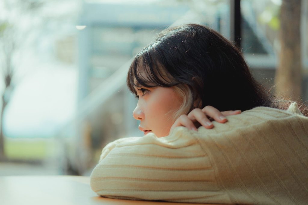 Young woman in a cream sweater rests her head on her arms beside a gray window, representing seasonal depression support through DBT therapy at DBT of South Jersey in Voorhees and Moorestown, NJ